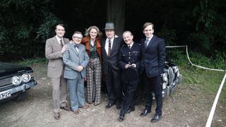 The cast of Endeavour - Sean Rigby, James Bradshaw, Abigail Thaw, Roger Allam, Anton Lesser and Shaun Evans - stand together beside the black Jaguar car.
