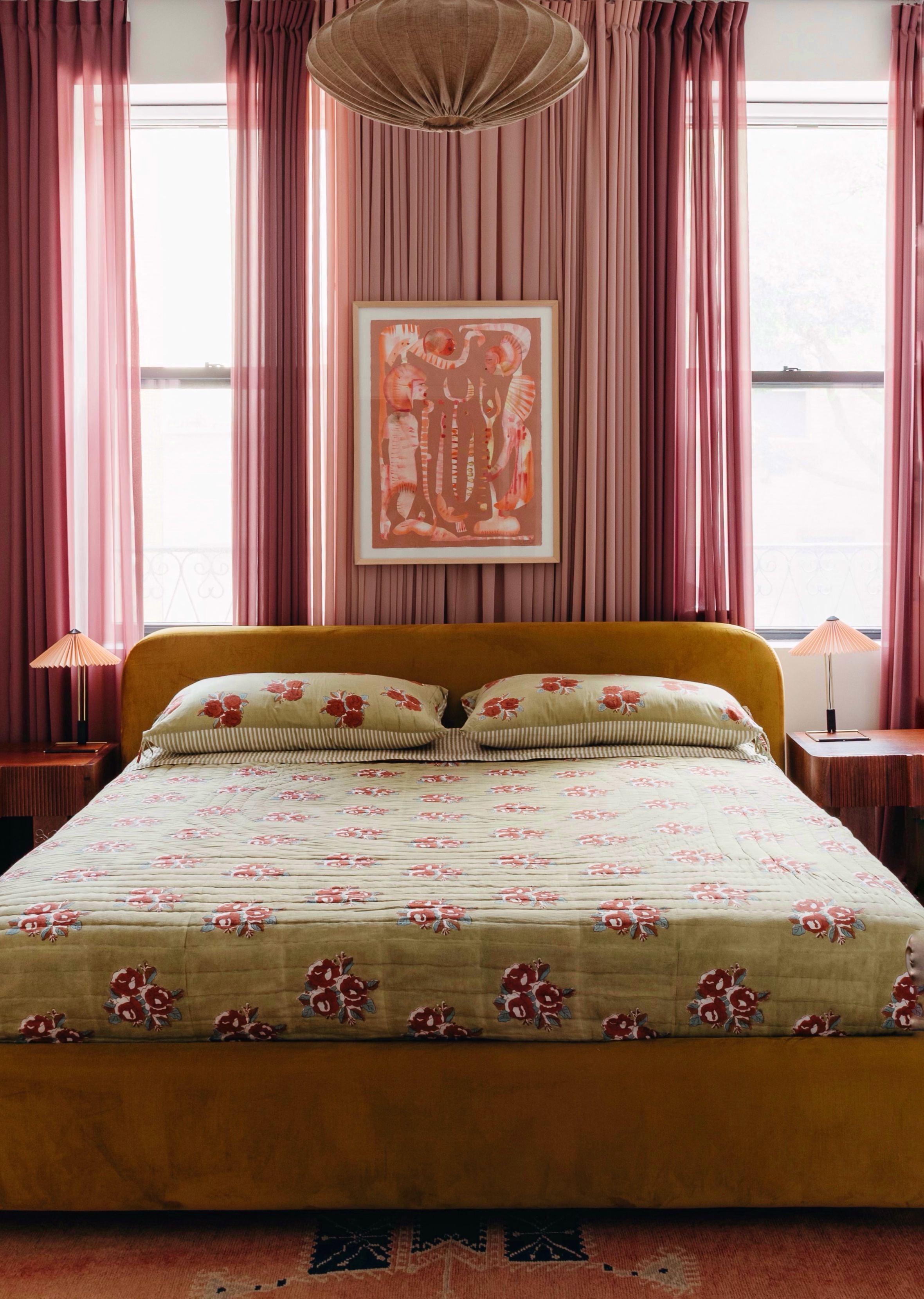 A green and pink toned bedroom where an artwork hangs above the headboard against heavy wall-to-wall drapes.