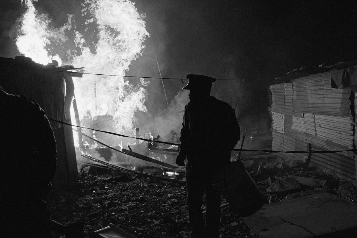 Black and white photograph of a man. Behind him there is a fire burning