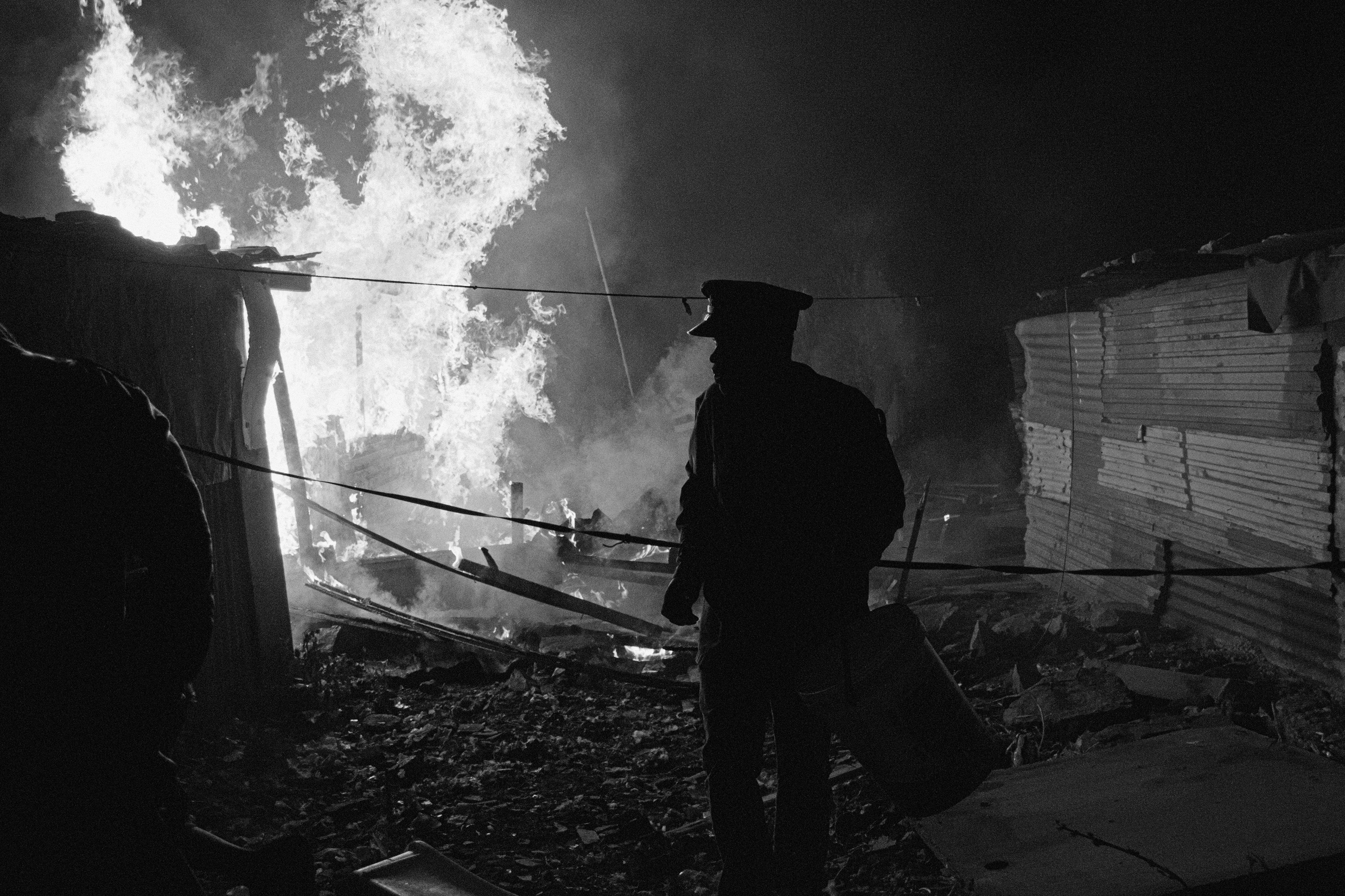 Black and white photograph of a man. Behind him there is a fire burning