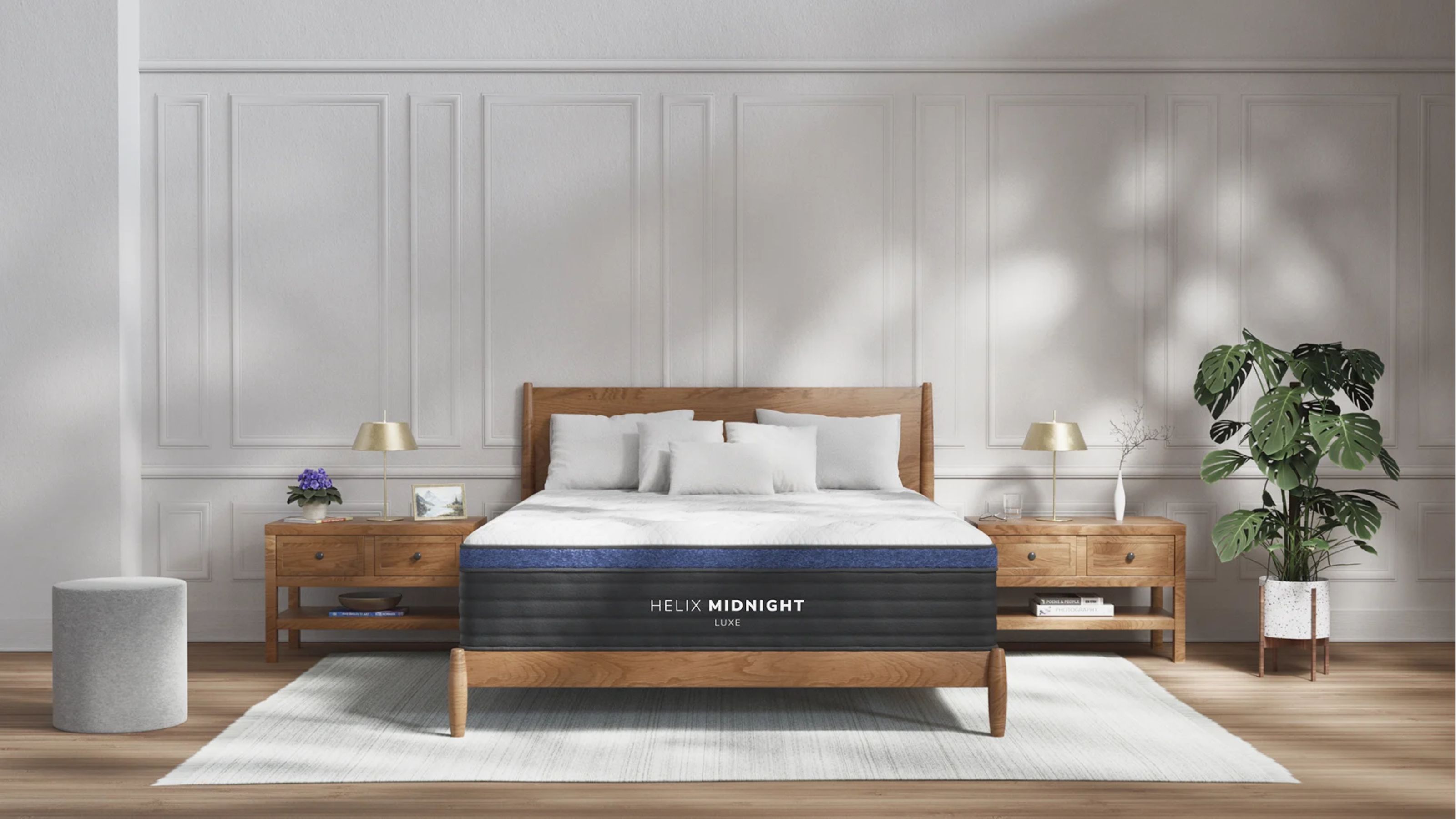 Helix Midnight Luxe Mattress pictured on a wooden bed frame in a large bedroom with white panelled walls, wooden flooring, a pale grey rug, and large houseplant, with two matching wooden nightstands.