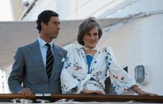 Prince Charles wearing a gray suit standing on the Royal Yacht Britannia next to Princess Diana, wearing a white floral print dress that ties in front and a pearl choker