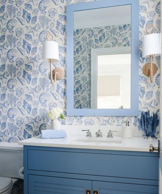 A coastal small bathroom with blue and white shell patterned wallpaper, a cornflower blue rectangular mirror, two wall sconces either side, and a vanity unit with blue cabnets and a white counter