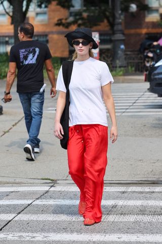 Jennifer Lawrence wearing a black hat, black sunglasses, a white t-shirt, red pants, and a black shoulder bag.