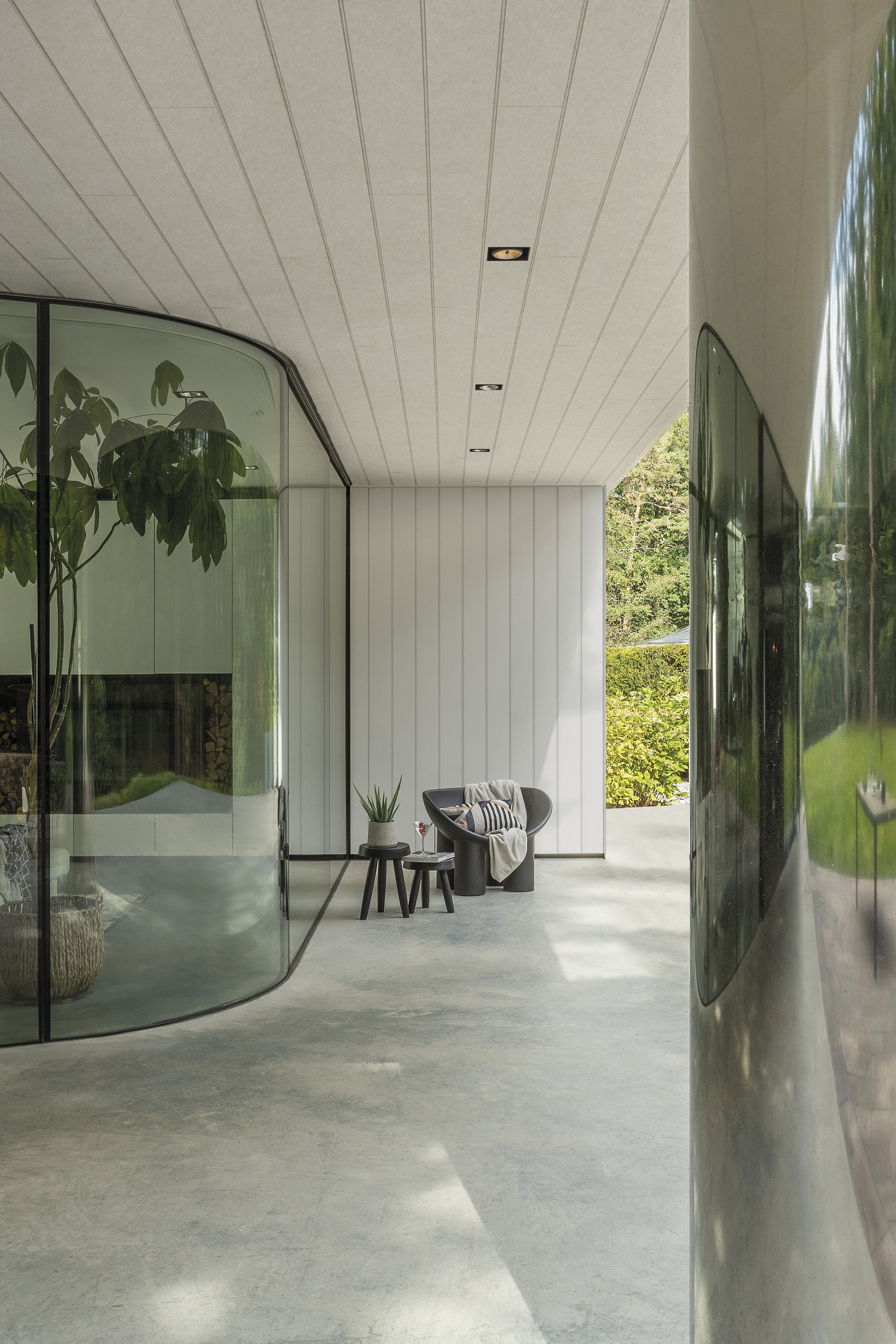 Image of a covered outdoor space where the ceiling and walls are covered in a white fibre cement cladding. The walls of the interior are made of a glass window, and there is a small chair and side table by the back wall.