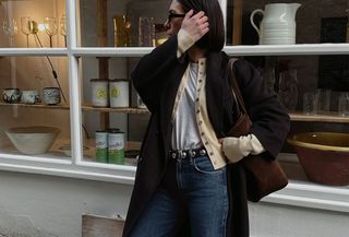 Woman wearing a black coat over a tan cardigan, white tee, and jeans.