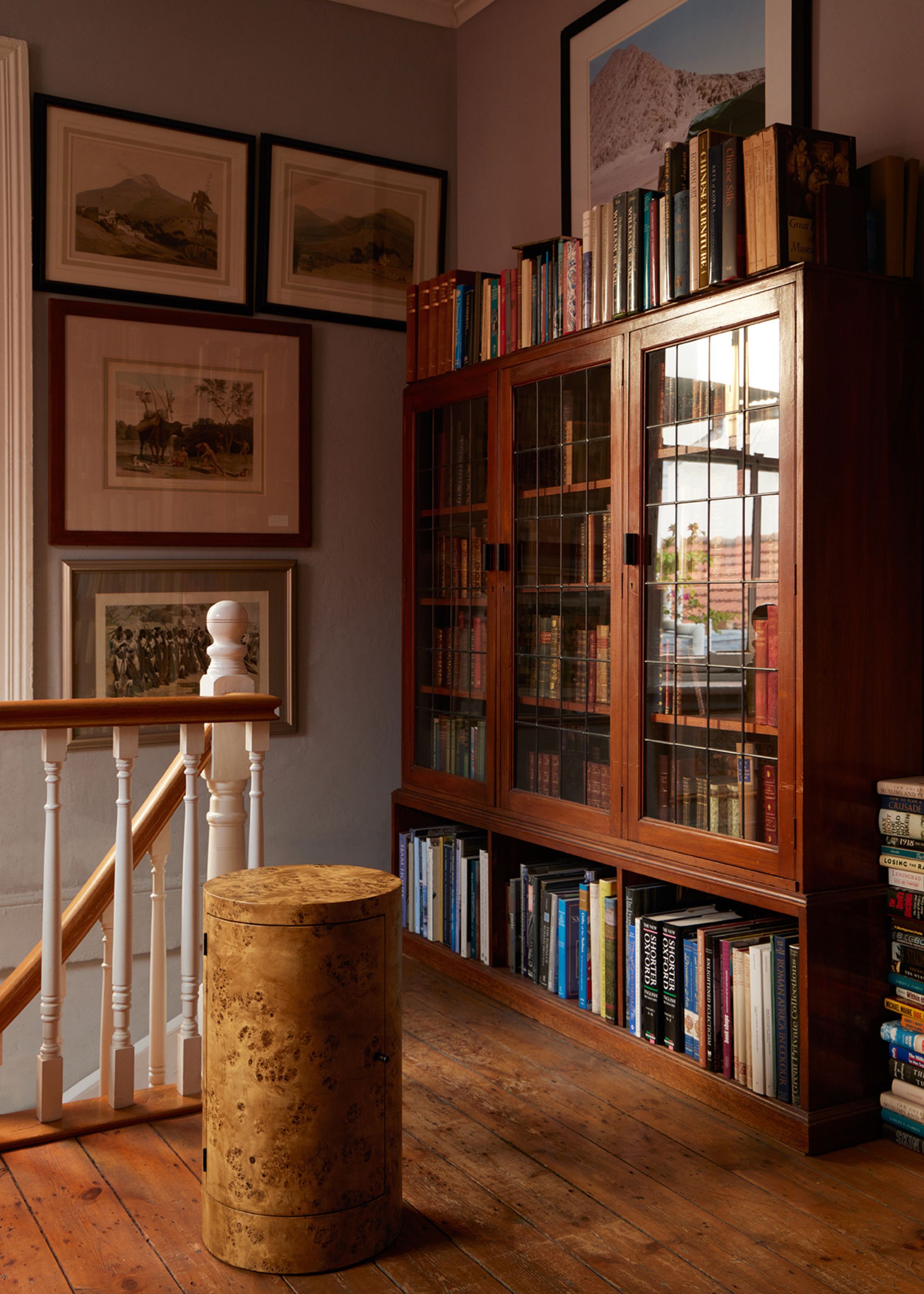 A home with a book case stacked with books of different sizes and colors, beside a burl wood seat, and a wall of landscape art