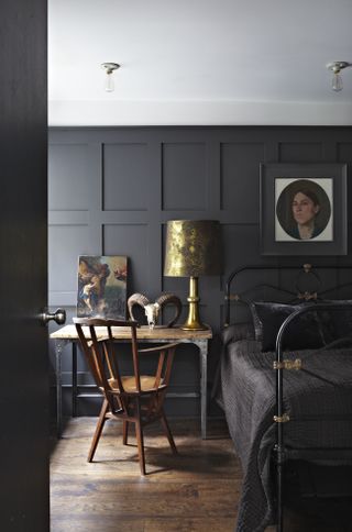 black bedroom with black panelled walls, black iron bed, vintage desk and chair, brass table lamp, wooden floors, white ceiling, artwork