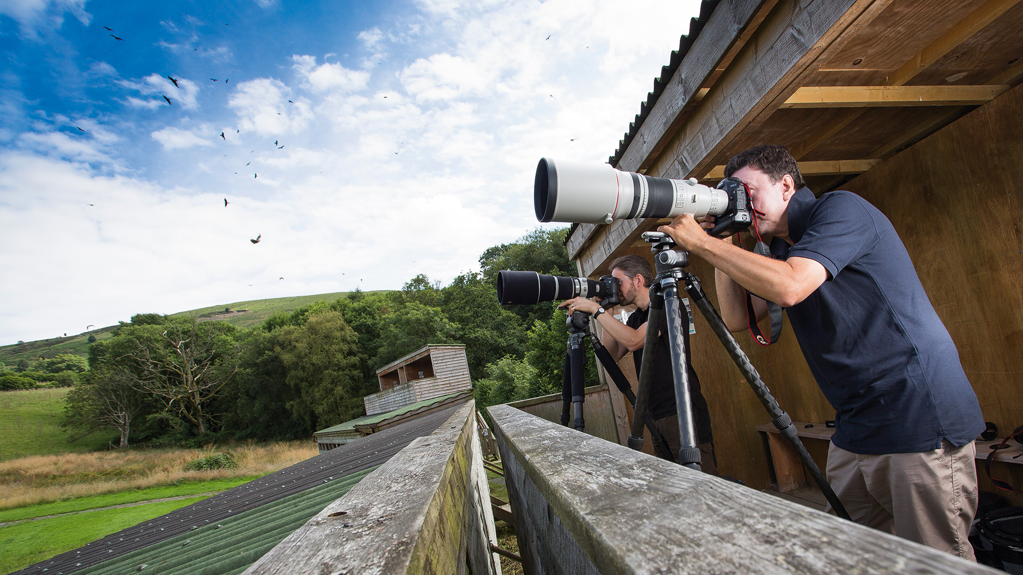 Two photographers taking photos from a hide against a blue sky with birds in the sky 