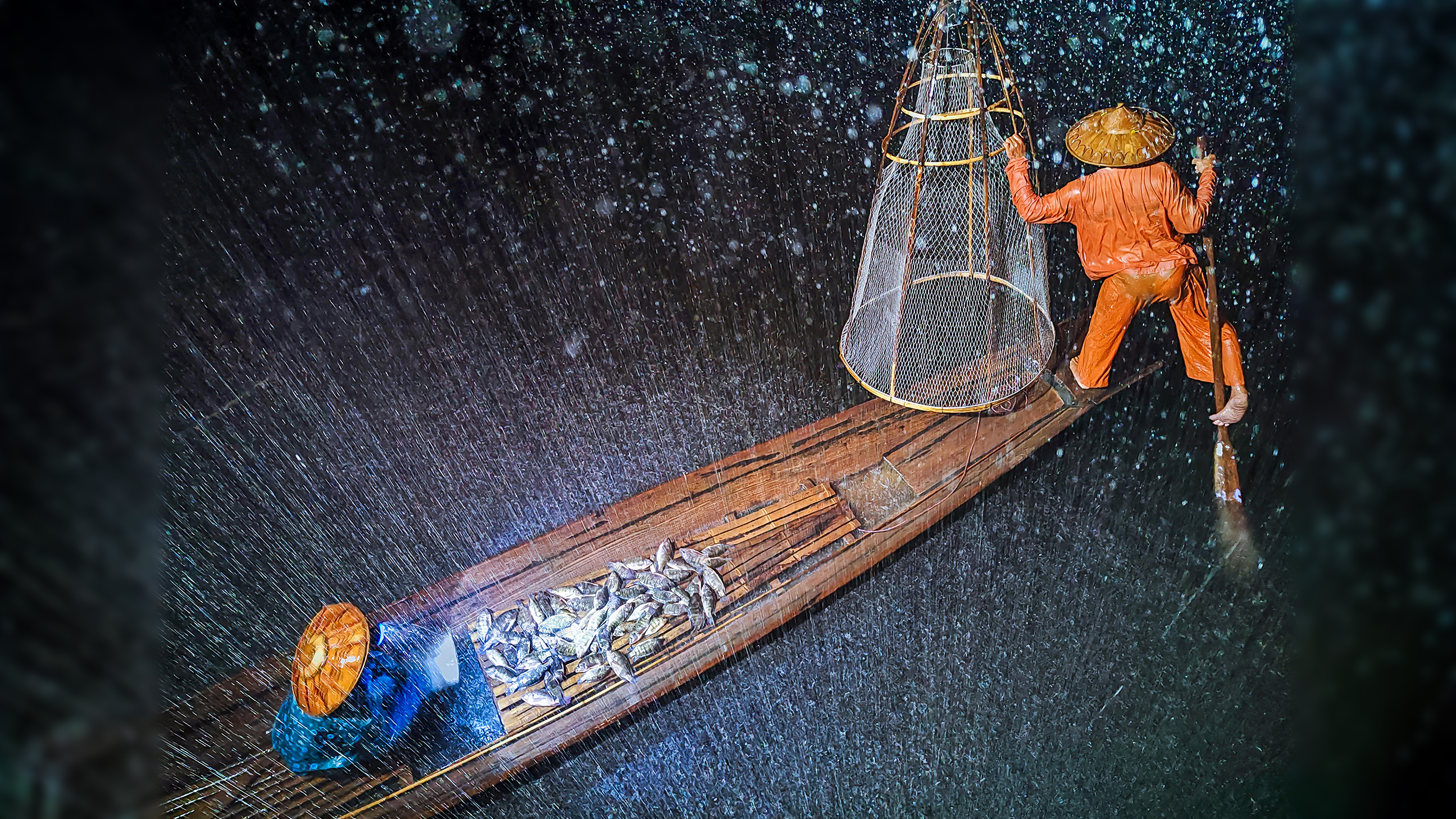 A fisherman in orange attire navigates a wooden boat, catching fish under heavy rain, with water splashing around him