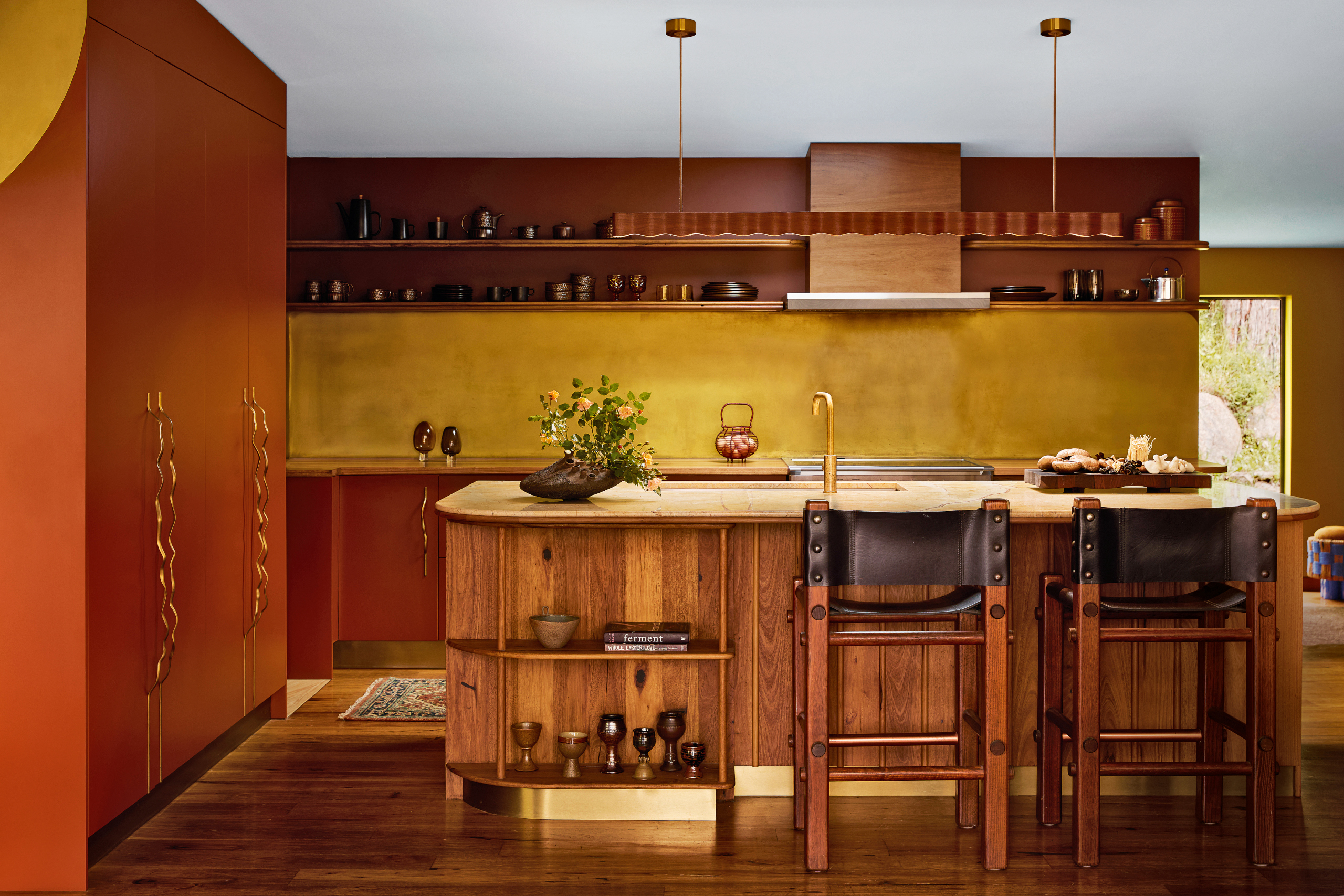 Kitchen with rust-red cabinetry, brass splashback and wood-panelled island with black leather and wood bar stools