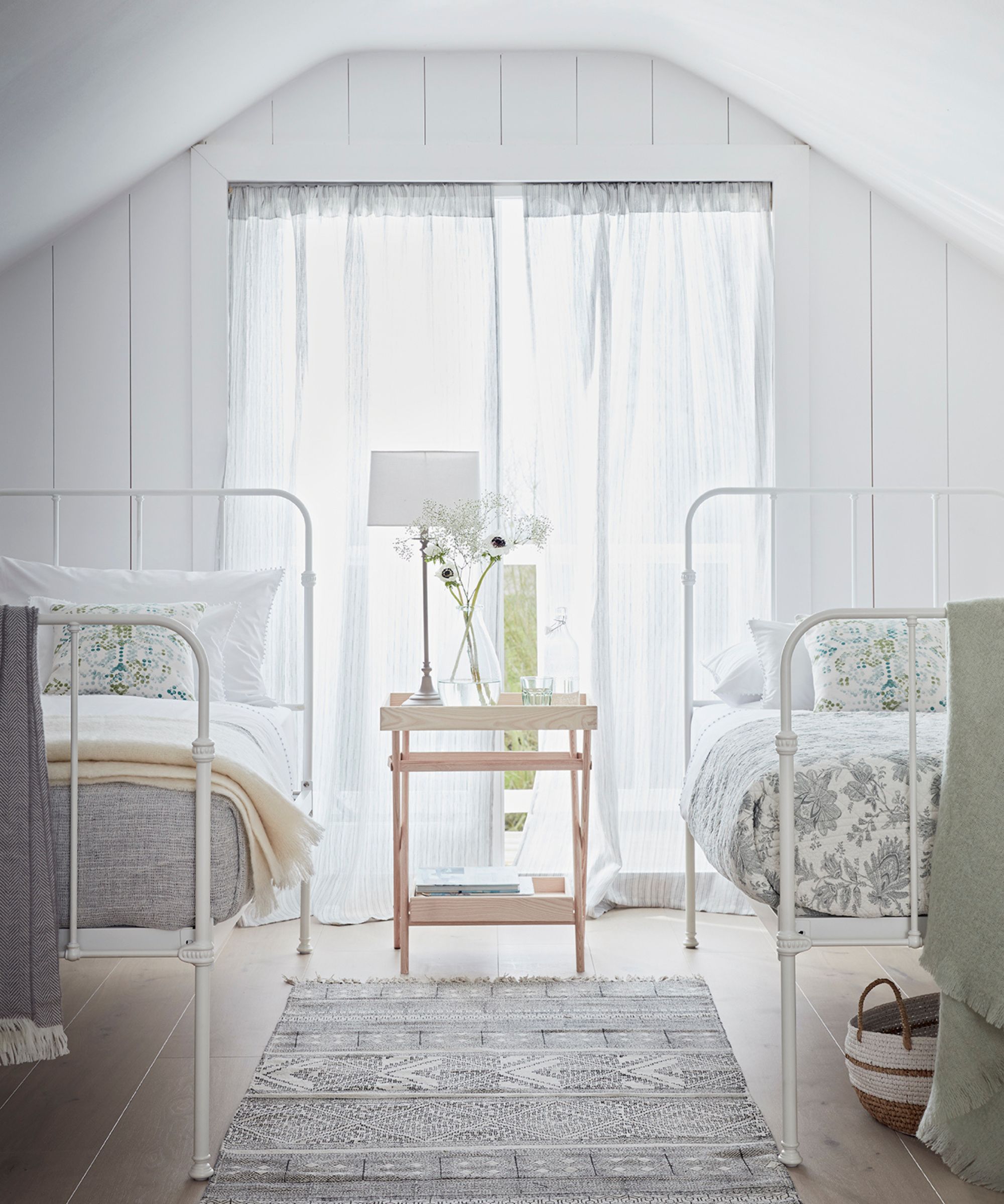 A bright, white twin bedroom in an attic with a large window with sheer white curtains, and a white panelled wall. In the centre of the room is a wooden bedside table with white lamp and glass vase of white flowers, and to either side are white metal beds with floral bedding, and a rug between. Beneath the right bed is a small white and brown storage basket. Hanging over the end of each bed is a blanket.