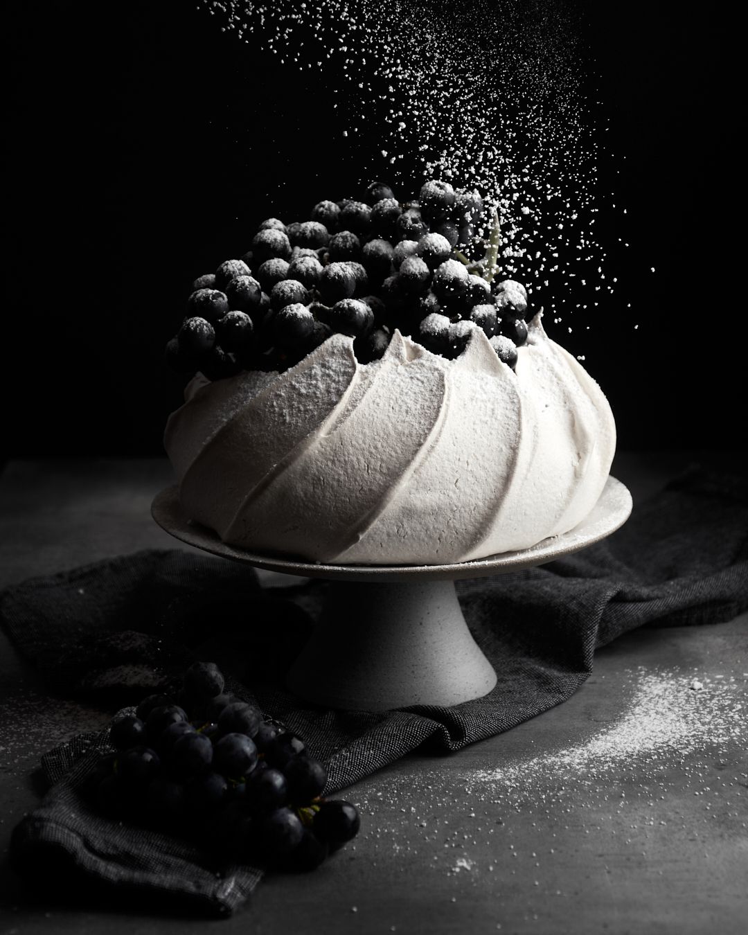 A beautifully styled pavlova topped with fresh blueberries, dusted with powdered sugar, set against a dark backdrop