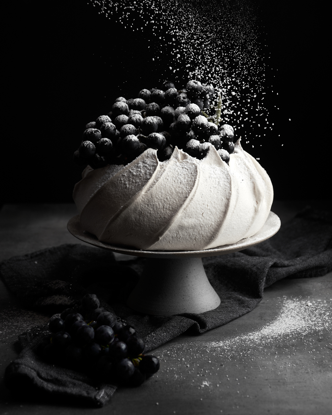 A beautifully styled pavlova topped with fresh blueberries, dusted with powdered sugar, set against a dark backdrop