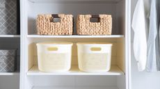 Organized white closet with multiple baskets on the shelves and several articles of clothing hanging from the closet rod