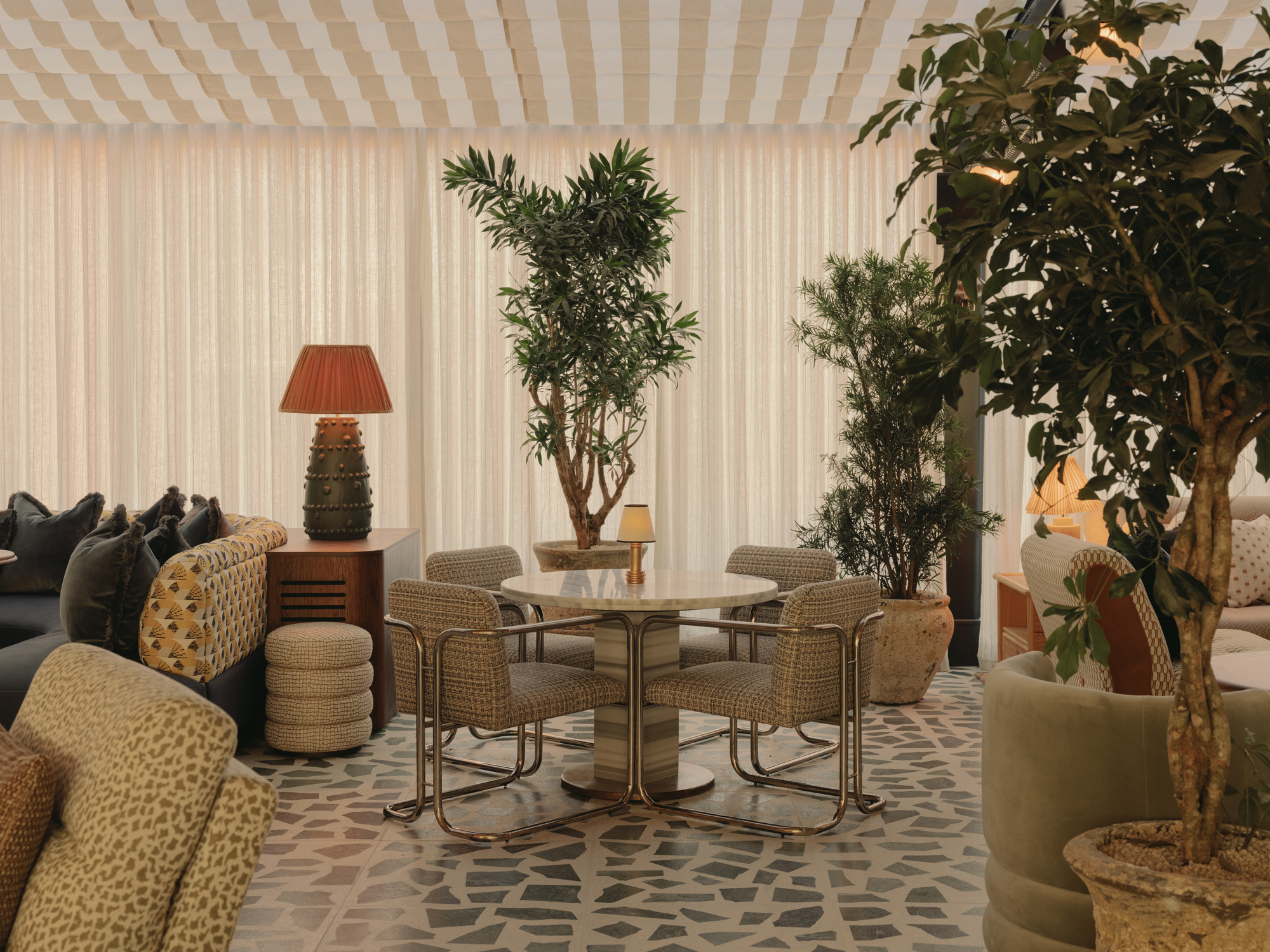 dining space in soho house manchester with striped canopy-style tented ceiling, round stone table with four wicker cantilevered chairs, potted trees, terrazzo floors, sofas with ottomans, a console with a table lamp and a wall of sheer curtains