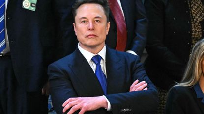 Elon Musk in a blue suit with his arms crossed over his body