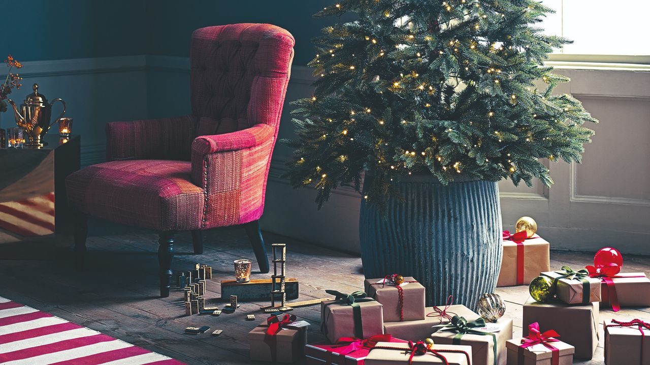 A Christmas tree in a planter with presents all around it and a red chair to the side