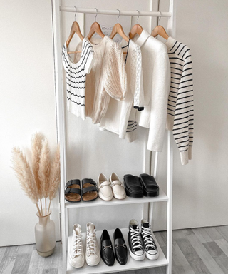 A white clothing rack with neutral fall clothing and knitwear next to pampas grass