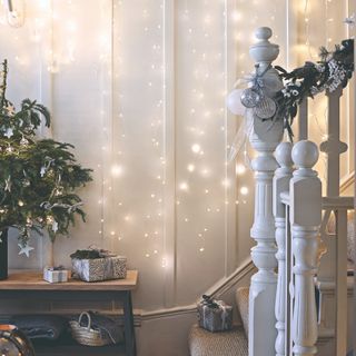 A white hallway decorated for Christmas with string lights hung on the panelled walls and a small Christmas tree on top of the shoe bench