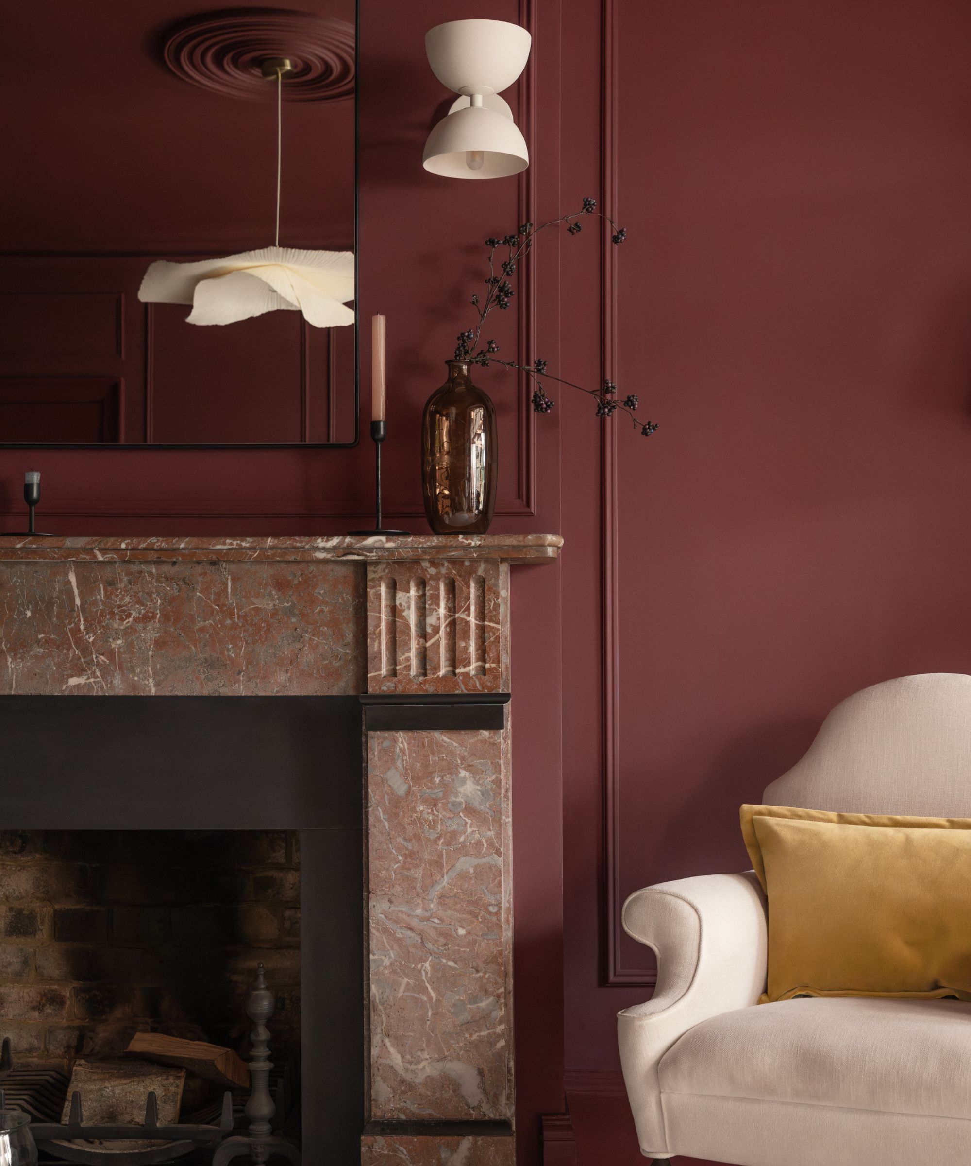 A living room with dark red walls, a marble fireplace with a mirror above it, and a beige armchair with a mustard cushion.