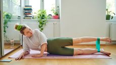 A woman performs a lying leg raise with a resistance band looped around her ankles. She is inside, on a yoga mat, lying on her side propped up on her elbow with her legs elongated. Her top leg is elevated so that it's parallel to the floor.