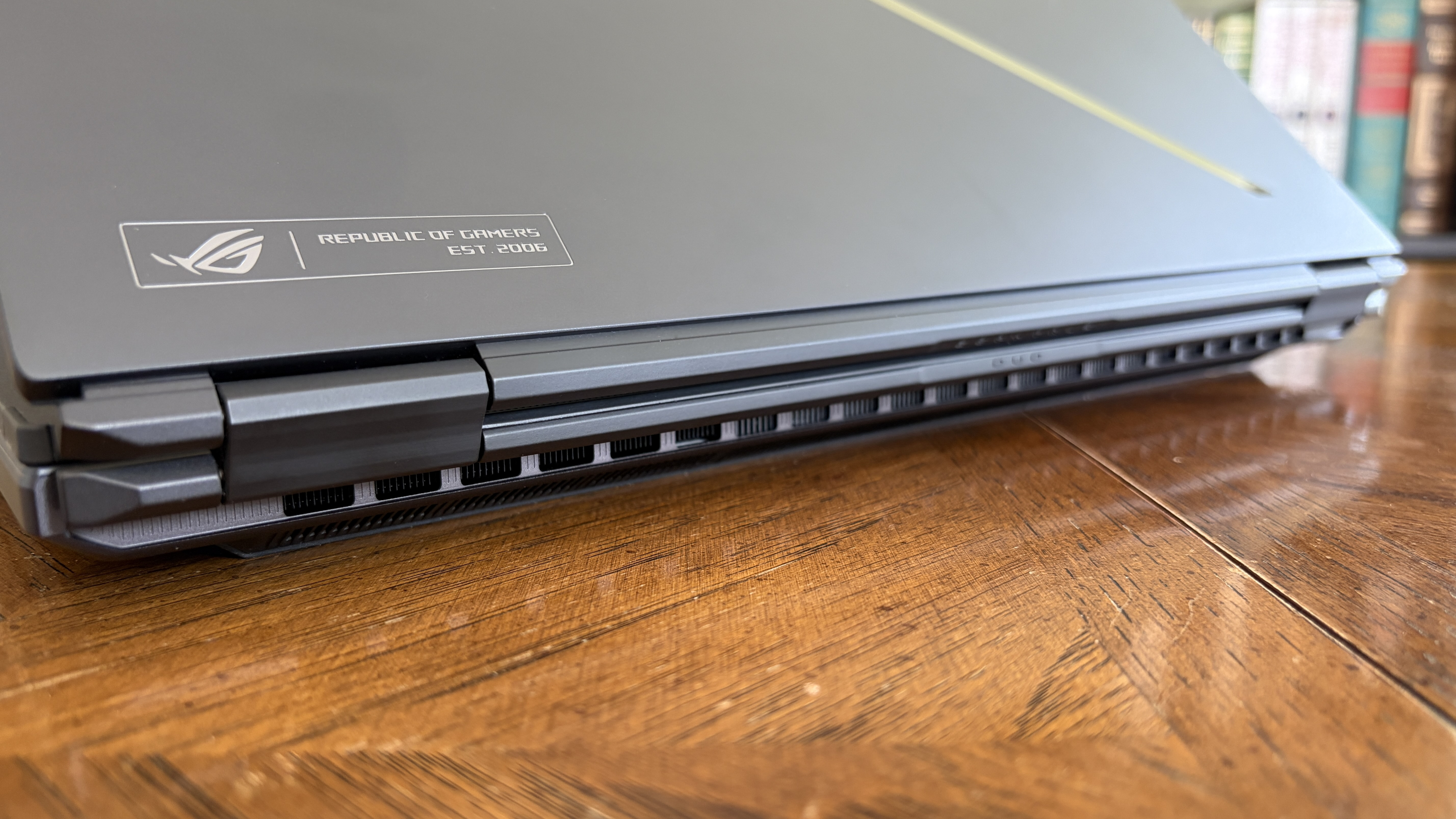 The image shows a closed gaming laptop with a &amp;quot;Republic of Gamers&amp;quot; logo on the lid. It's placed on a wooden surface, with books blurred in the background.