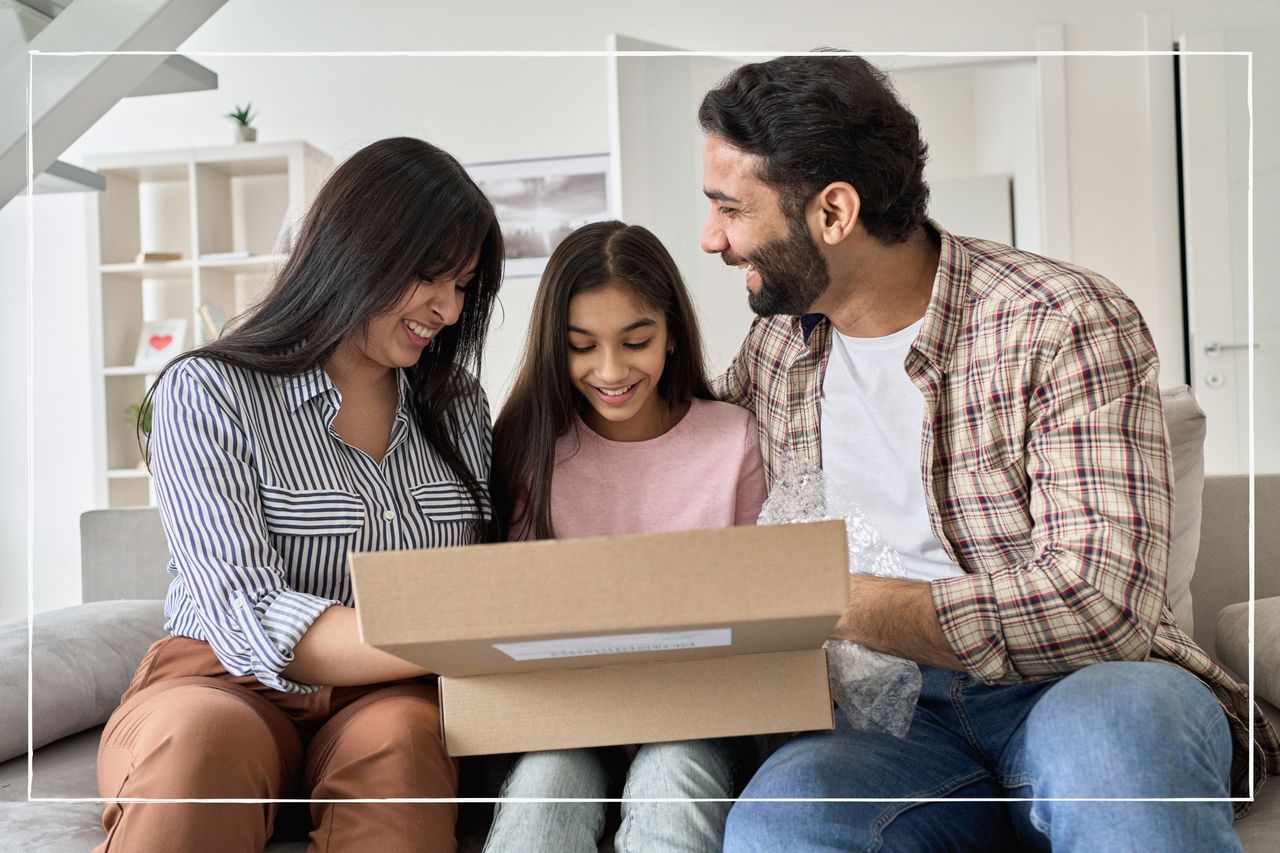 Parents and daughter smiling as they open a parcel together, while sitting on a sofa at home