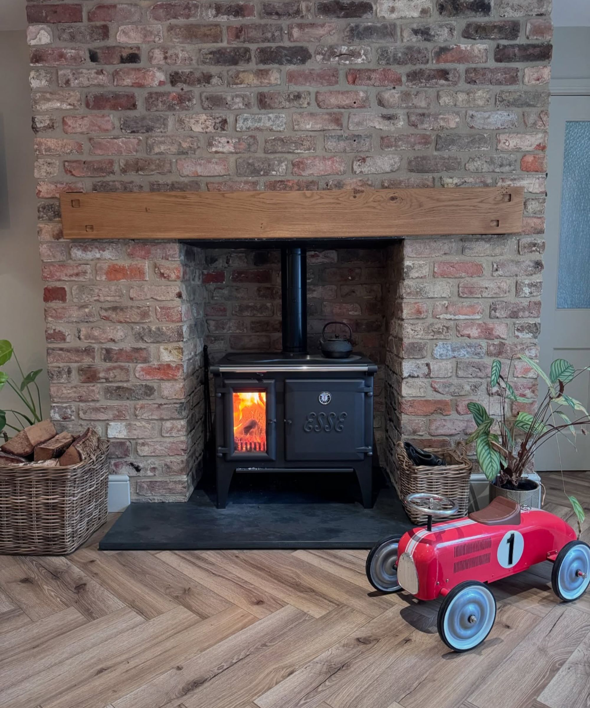 A rustic exposed brick fireplace with a black cast-iron log burner beneath a solid oak mantel beam. A wicker basket of logs sits nearby, and a child&amp;rsquo;s red vintage-style toy car rests on the wooden herringbone floor, adding a playful, homely touch.