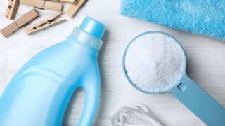 A bottle of liquid laundry detergent next to a scoop filled with powder laundry detergent next to some clothes pegs and towels