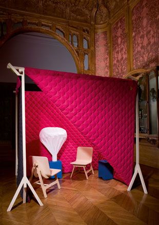 Interior room, wooden floor, red patterned walls, red textured screen on white stand, wooden archway, white balloon shaped feature on a blue box, two wooden chairs