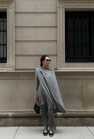 a photo showing the fall jacket trend, capes worn by a woman standing against a tan wall wearing a light gray knit cape layered over a matching gray top, knit pants, black ballet flats, and a black bag