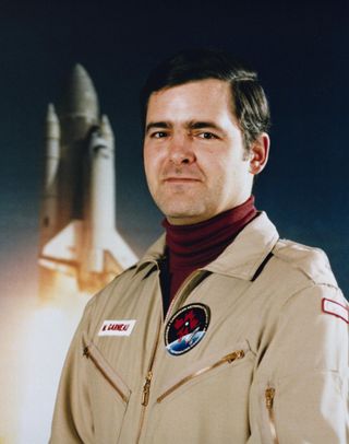 marc garneau in a tan flight suit in front of a picture of a space shuttle lifting off