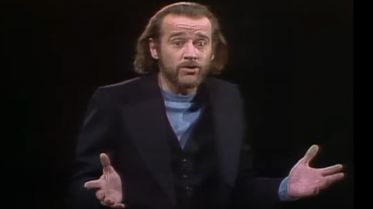 George Carlin with long hair, handlebar mustache, performing while hosting the first episode of Saturday Night Live.