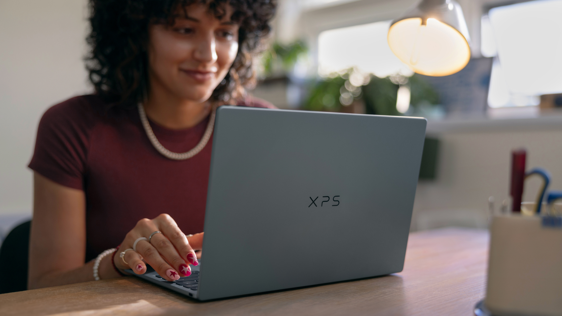 Person sitting at a desk using a Dell XPS laptop, with a lamp, plants, and office supplies in the background.