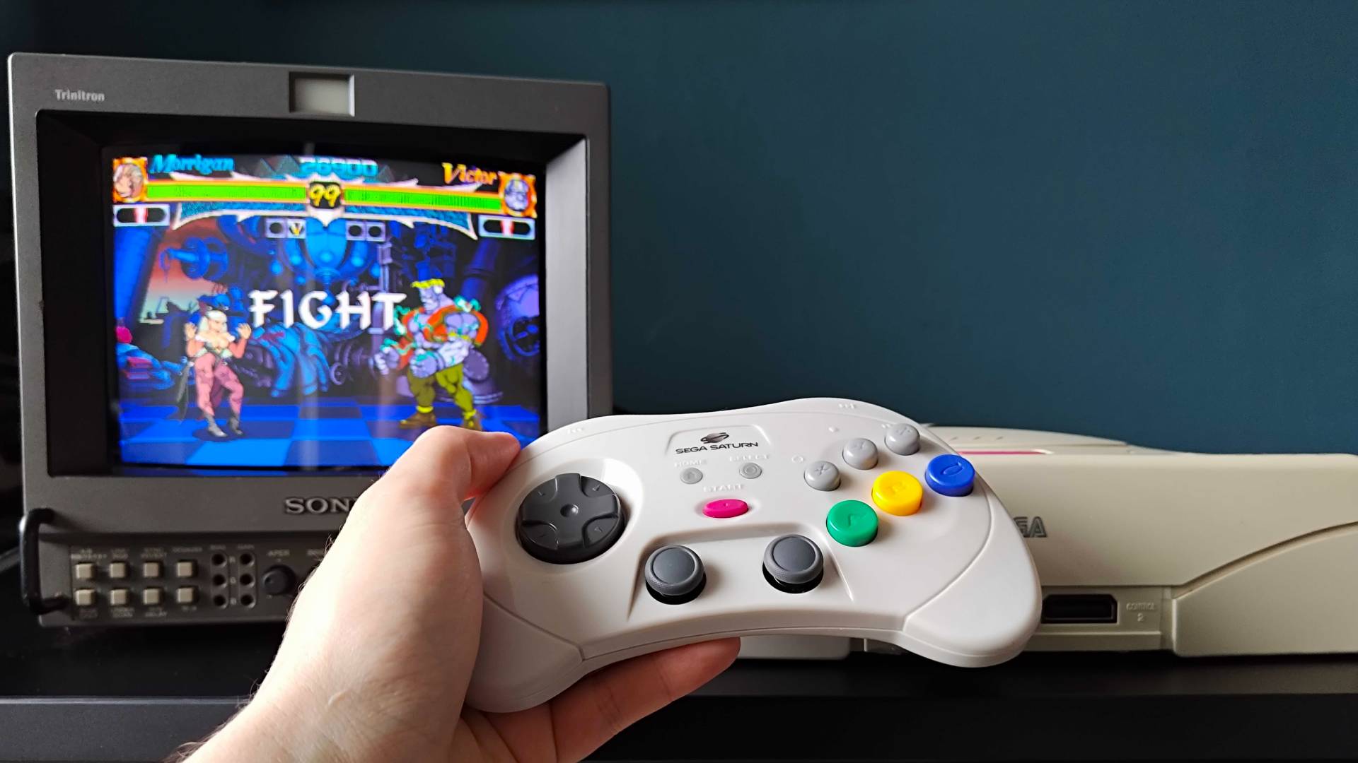 Hand holding Retro-Bit Sega Saturn Pro controller in front of matching white console and Sony PVM CRT TV with Vampire Hunter Darkstalkers Revenge gameplay on screen.