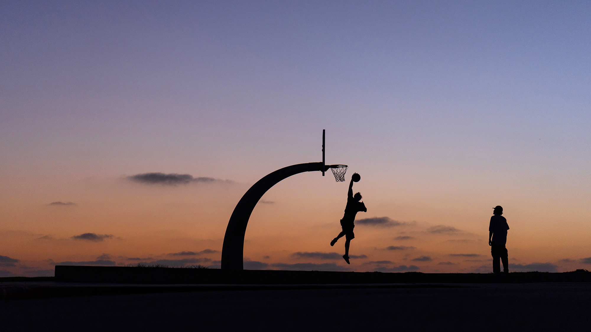Two people are silhouetted playing basketball at dusk in the San Pedro neighborhood of Los Angeles