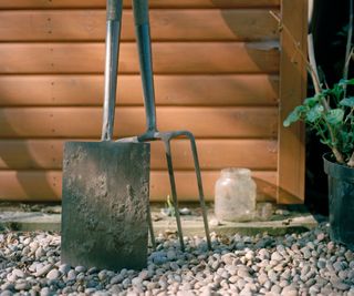 A muddy spade in front of a garden fork by a shed