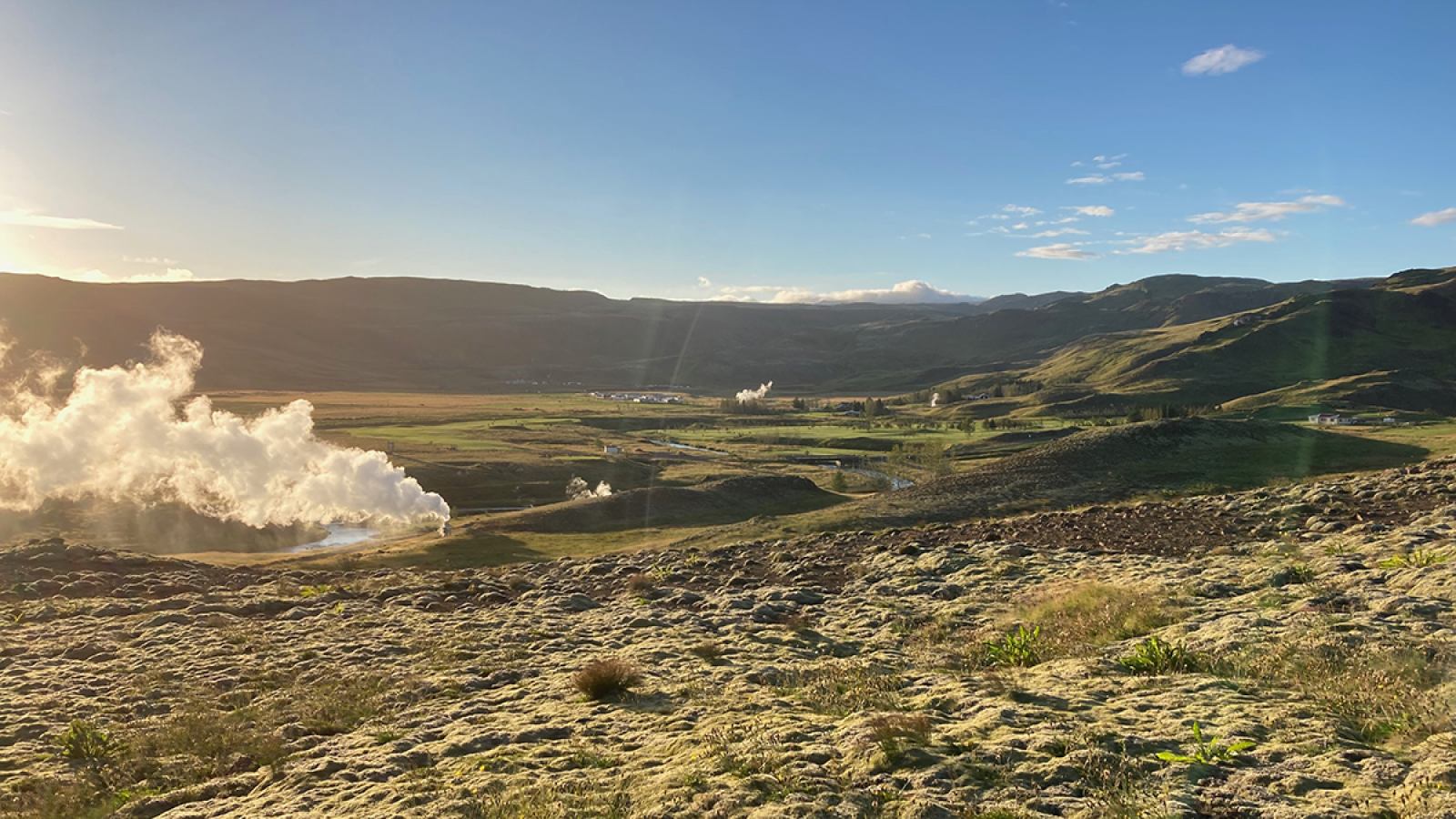 Geothermal activity, with steam rising over the grassy ground and rolling hills