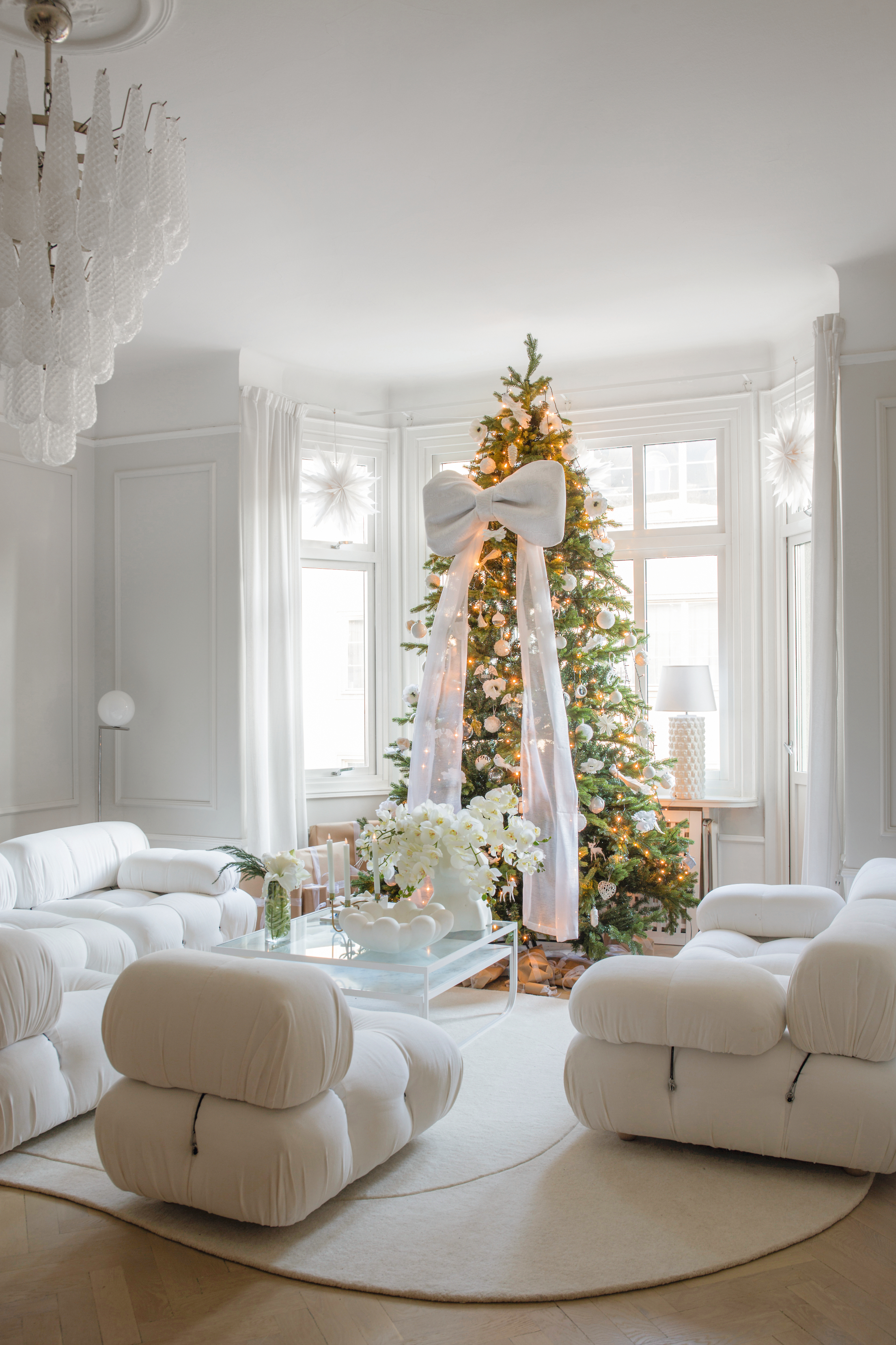A white living room with white B&amp;amp;B Italia sofa, glass coffee table and Christmas tree with oversized white bow
