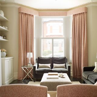 A beige living room with salmon pink curtains containing three sofas arranged around an upholstered coffee table