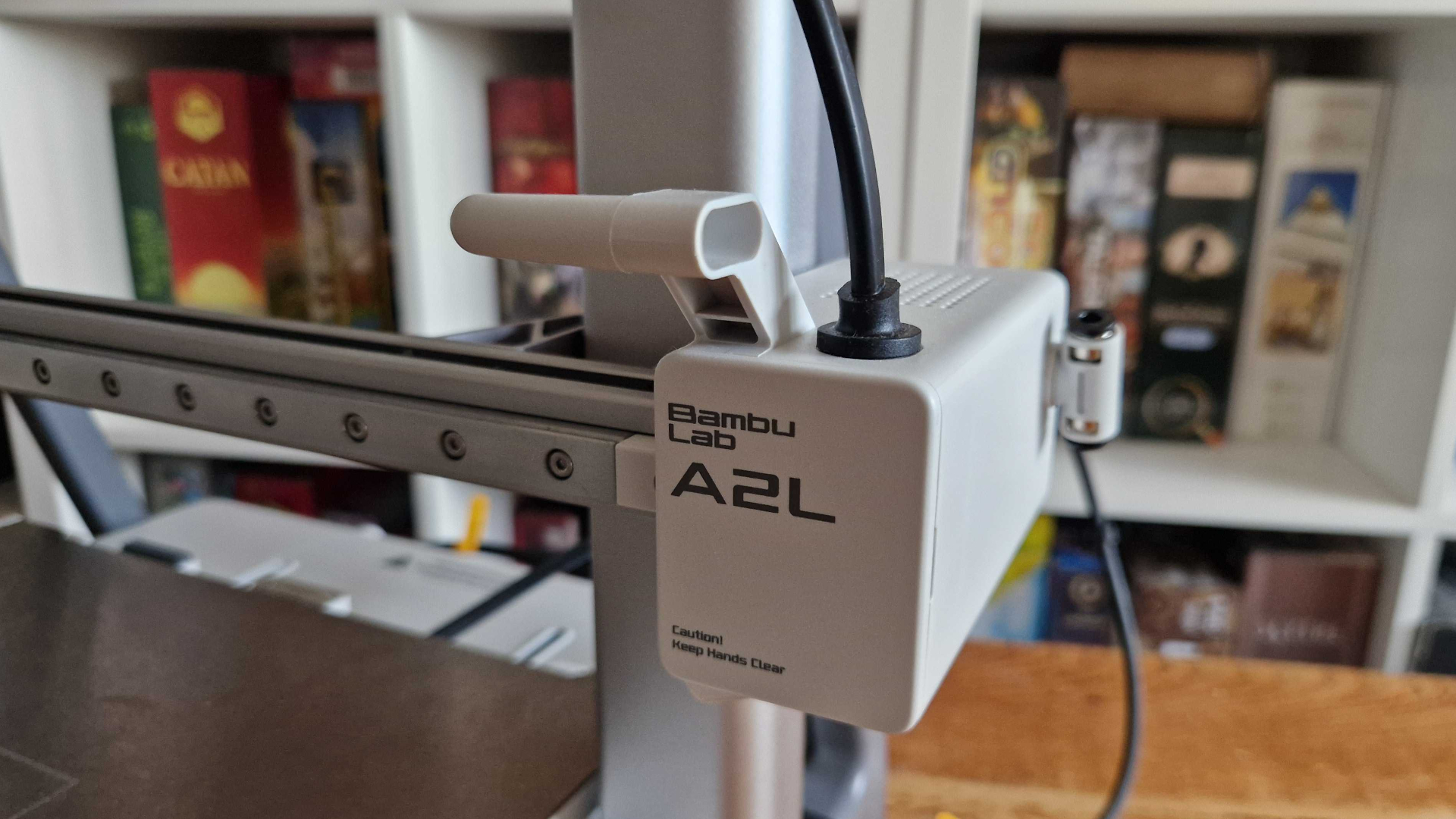 The Bambu Lab A2L logo on the machine itself, with board game boxes visible in the background