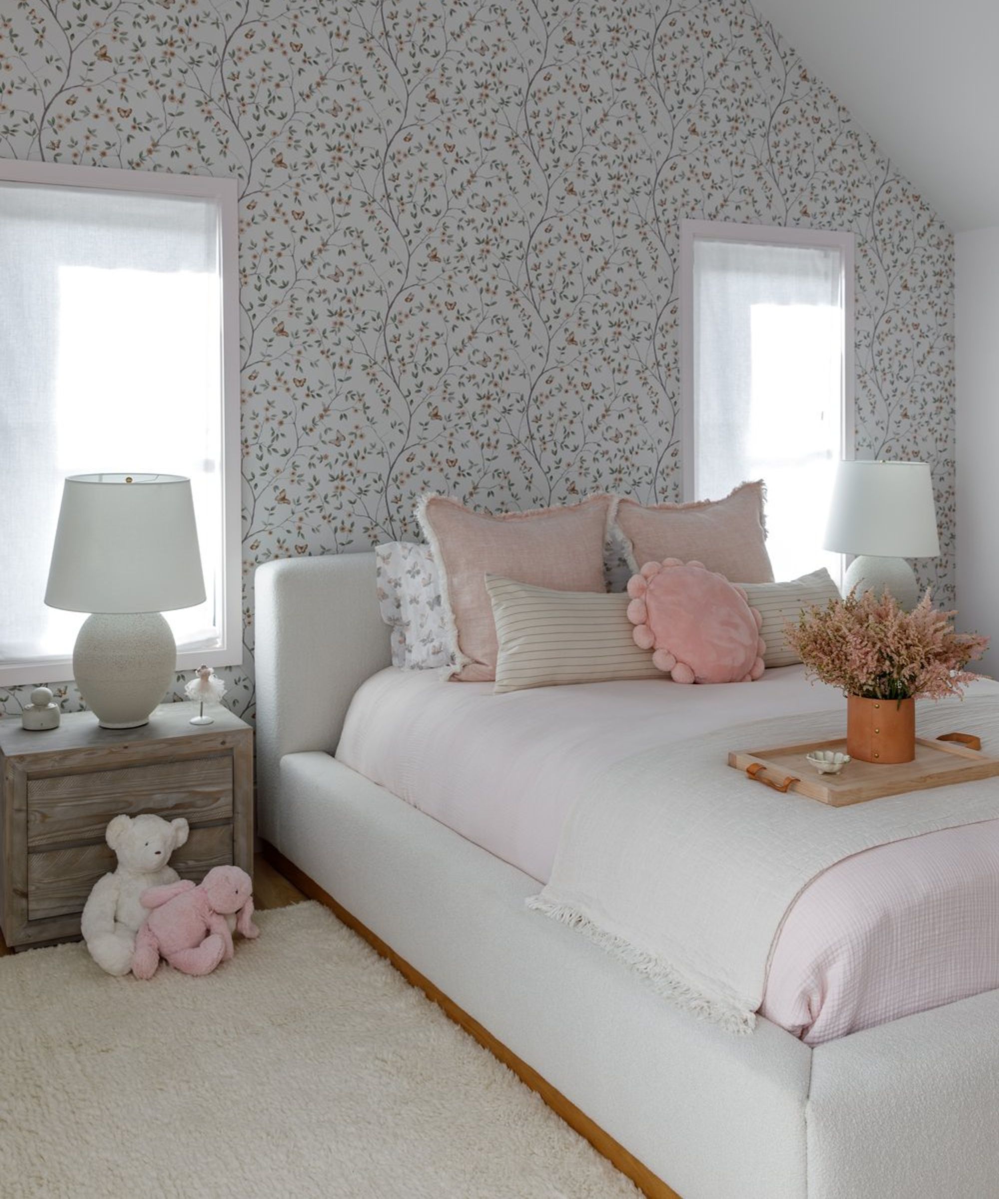 Floral wallpaper on bedroom wall with two windows, and pitched ceiling, with tightly tucked in bedsheets
