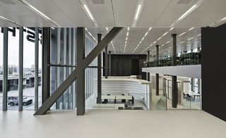 Interior view of the lobby at G-Star RAW headquarters, Amsterdam, white floor, white curved divider wall with black seating on the ground floor, grey pillars and beams, white tiled ceiling with strobe lighting, ground floor and upper floor desks and open plan office space, tall glass windows with view of surrounding area, car park and parked jeep, grey sky