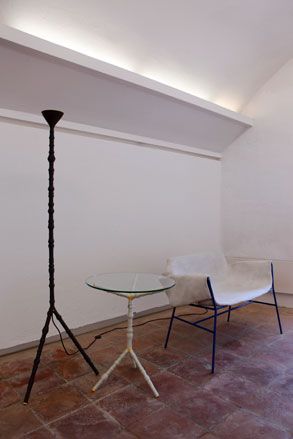 Black floor lamp next to a side glass table and chair, in a room with white walls and tiled brown concrete floor