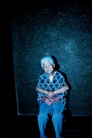 An older Japanese woman wearing blue leans against the wall with another pair of hands around her waist