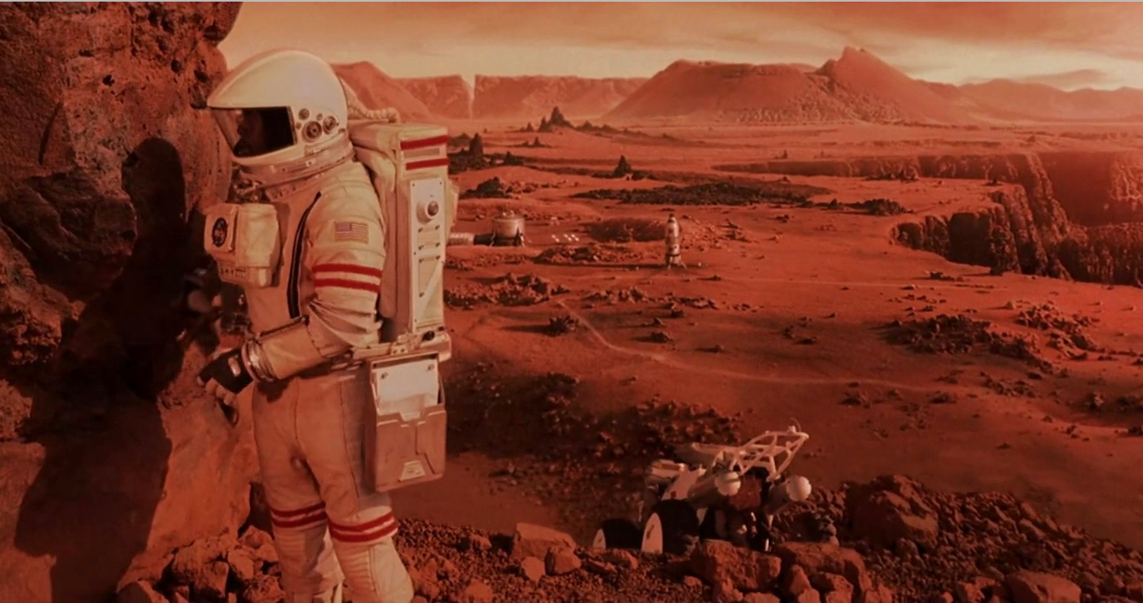 A lone astronaut stands beside a rock outcropping on a reddish planet