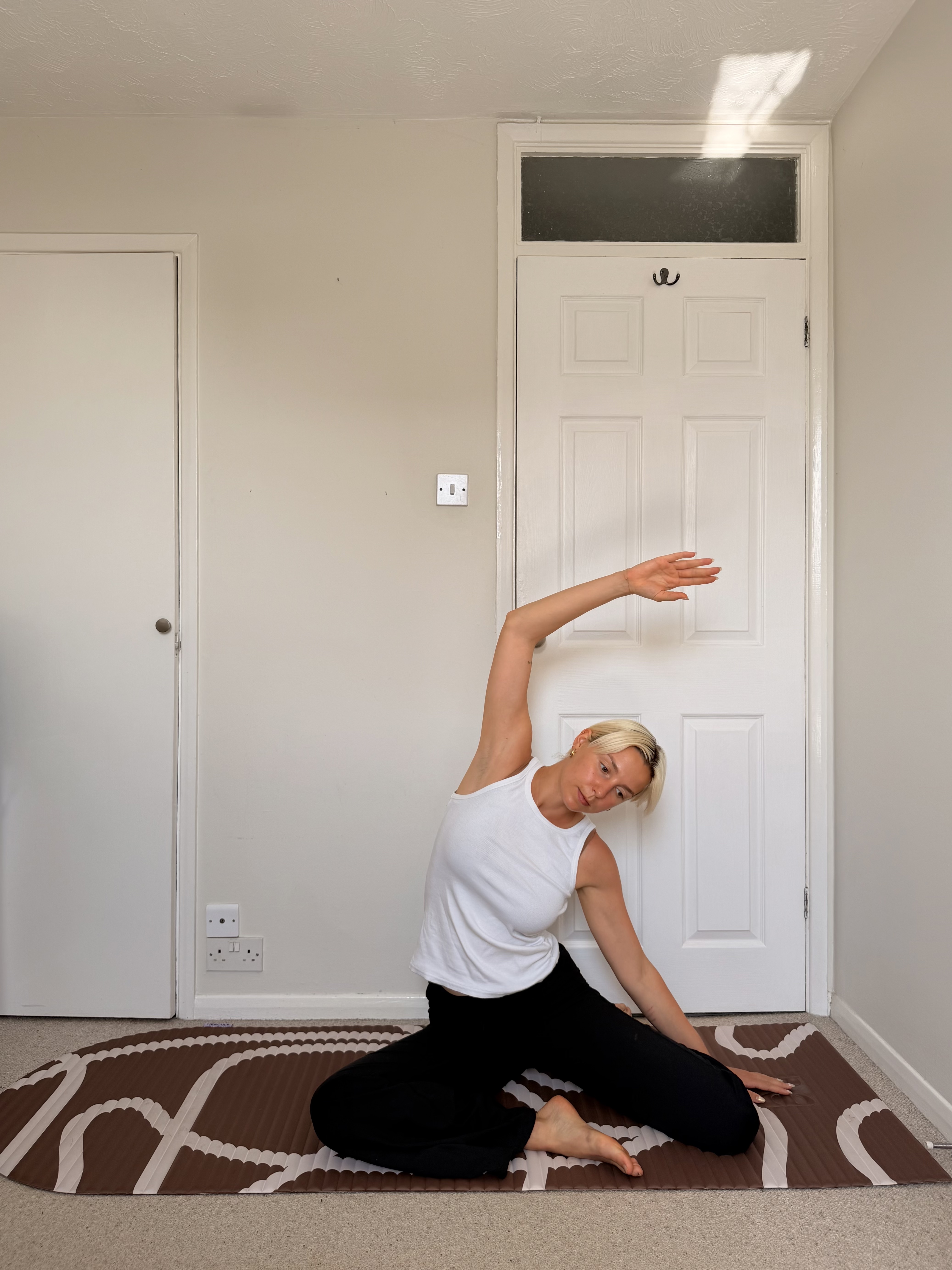 A woman in a white vest top and black trousers sitting on a brown yoga mat practicing a side stretch with her right arm raised overhead