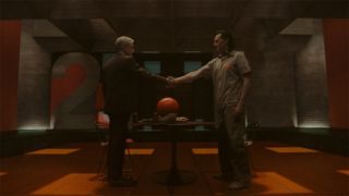 Image from the Marvel T.V. show Loki, season 2 episode 6. Zoomed out shot of two men shaking hands respectfully in an orange and gray room, a table between them. The man on the left has short white hair and is wearing a suit. The man on the right has slicked back dark hair and is wearing a prison-like gray jumpsuit.