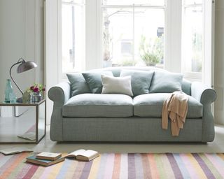 A grey upholstered sofa bed in living room with striped multicolored rug, metal side table and lamp light